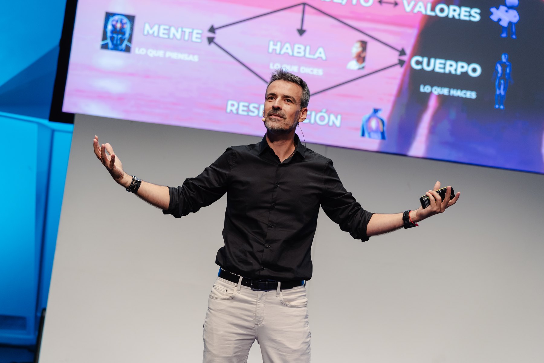 Antoni Fernández con los brazos abiertos en el escenario, ante una pantalla gigante con el triángulo del equilibrio: mente, habla, cuerpo y respiración
